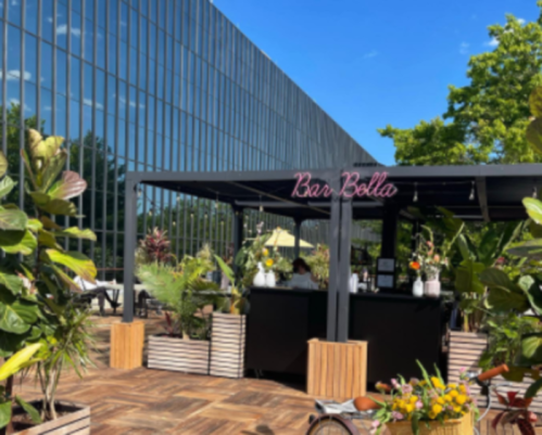 Outdoor bar area with a sign reading "Bar Bella," surrounded by plants and modern architecture under a clear blue sky.