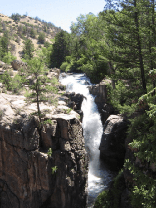A waterfall cascades through rocky cliffs, surrounded by lush greenery and trees under a clear blue sky.