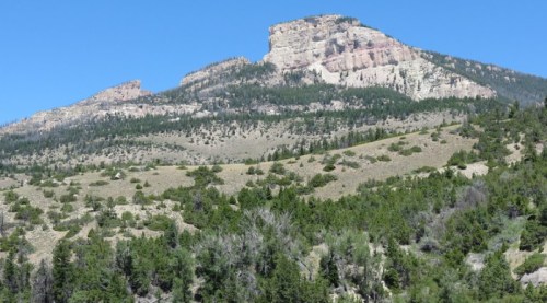 A rugged mountain peak rises above a green, forested hillside under a clear blue sky.