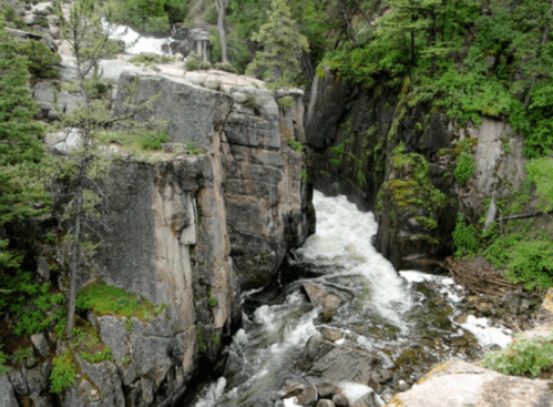 A rushing river flows through a rocky canyon surrounded by lush green trees and moss-covered boulders.