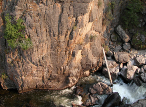 A rocky cliffside with greenery, overlooking a rushing river and large boulders below.