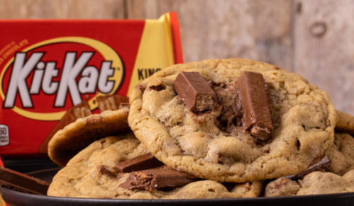A plate of cookies with Kit Kat pieces, next to a red Kit Kat package on a wooden background.