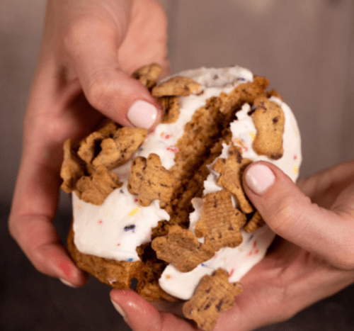 A person’s hands breaking apart a cookie sandwich filled with ice cream and topped with cookie pieces.