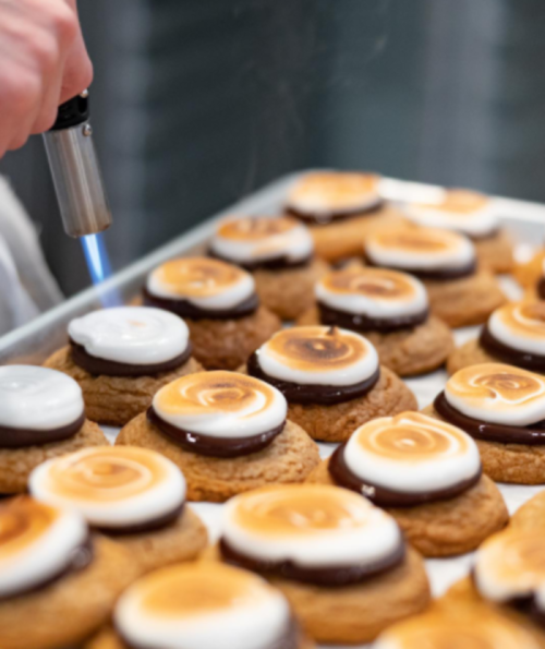 A hand uses a torch to caramelize the tops of cookies with chocolate and marshmallow frosting on a baking tray.