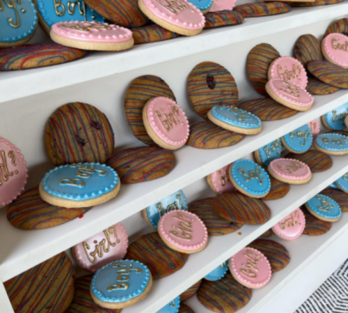 A display of colorful cookies with "Boy?" and "Girl?" written in gold, arranged on a white shelf.