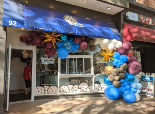 A storefront decorated with colorful balloons and a blue awning, featuring a sign for a cookie shop.
