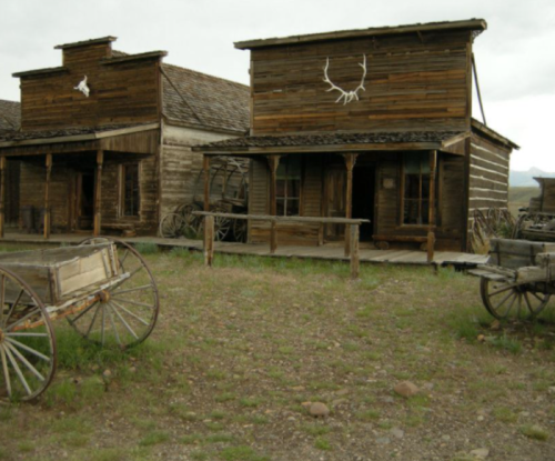 A rustic old western town with wooden buildings and vintage wagons in a grassy area under a cloudy sky.