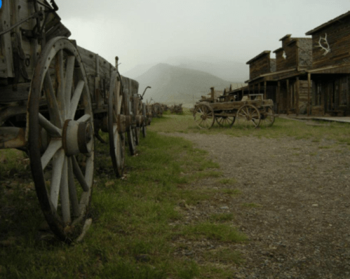 Old wooden wagons line a gravel path in a deserted western town, with misty mountains in the background.