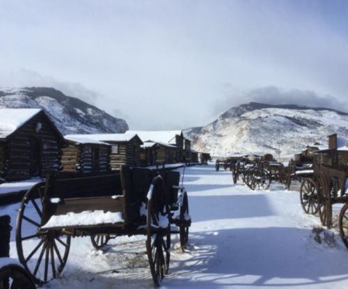 Snow-covered landscape with wooden wagons and rustic buildings, surrounded by mountains under a cloudy sky.