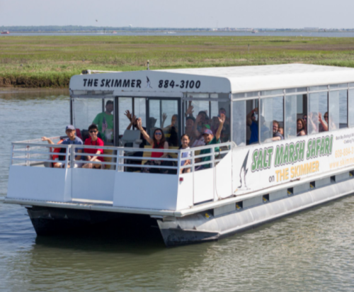 A group of people wave from a tour boat named "The Skimmer" on a scenic waterway.