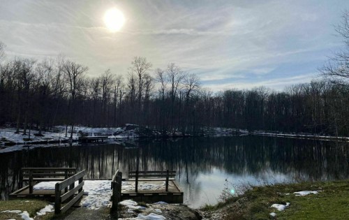 A serene lake surrounded by bare trees, with a bright sun overhead and patches of snow on the ground.