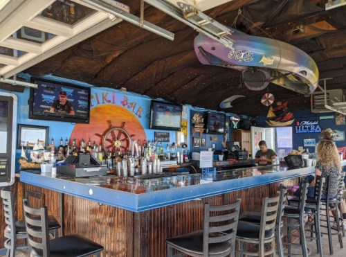 A tiki bar interior with a colorful mural, bar counter, bottles, and TVs, featuring a relaxed atmosphere.