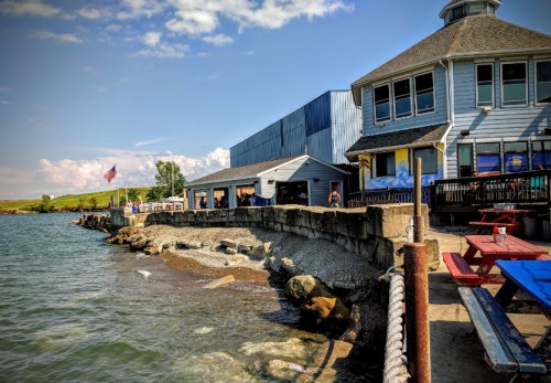 A waterfront view of a blue building with outdoor seating, rocky shore, and an American flag in the background.