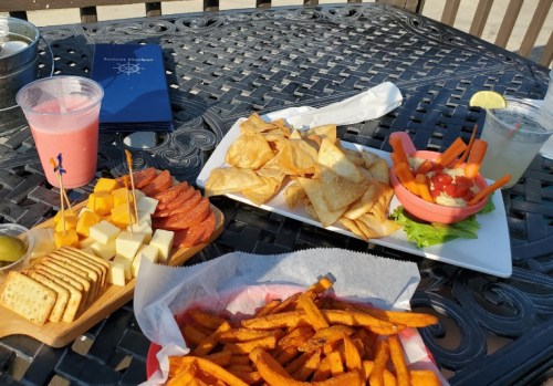 A table with a variety of snacks: sweet potato fries, cheese, crackers, chips, salsa, and drinks in a sunny outdoor setting.