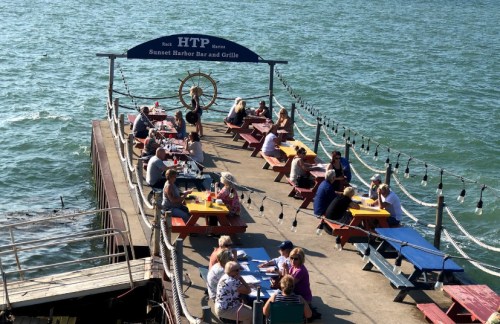A busy dockside bar and grill with colorful picnic tables, people dining, and a scenic view of the water.