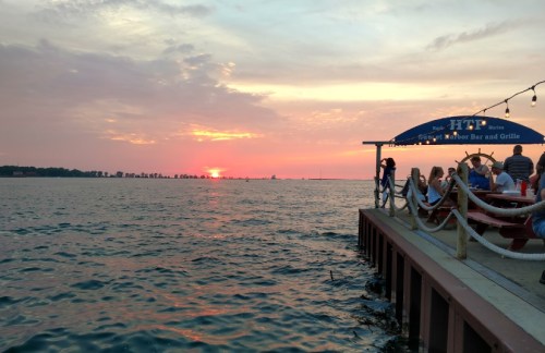 Sunset over water with a dock and people dining at a waterfront bar, surrounded by a colorful sky.