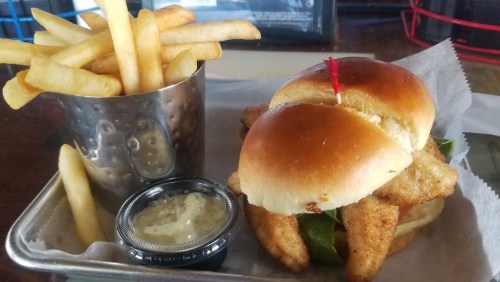 A plate featuring a sandwich with fried fish and lettuce, accompanied by a cup of crispy French fries and dipping sauce.