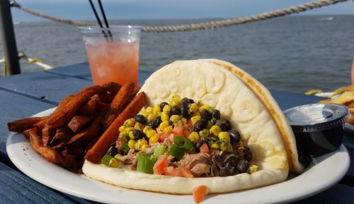A plate with a taco filled with meat, corn, black beans, and tomatoes, alongside sweet potato fries and a drink.