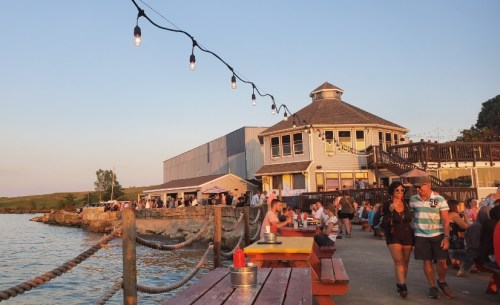 A waterfront scene at sunset with people walking, dining, and string lights illuminating a restaurant by the water.