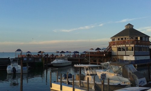 A waterfront scene featuring boats docked near a restaurant with outdoor seating and umbrellas under a clear sky.