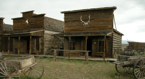 A rustic old western town with wooden buildings and vintage wagons on a grassy landscape under a cloudy sky.
