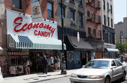 A street view of Economy Candy shop with people outside, surrounded by urban buildings and parked cars.