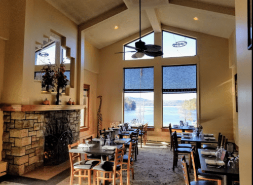 Cozy restaurant interior with stone fireplace, tables set for dining, and large windows overlooking a scenic lake view.