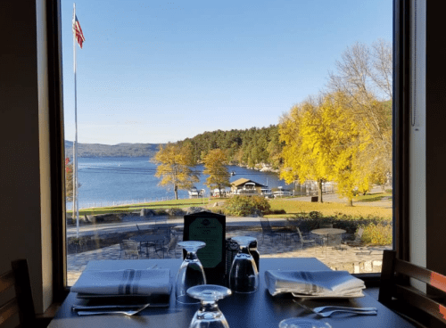 A scenic view of a lake and autumn trees from a restaurant window, with a table set for dining in the foreground.