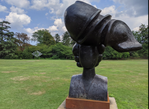 A large abstract sculpture of a head with a helmet, set in a grassy park with trees and clouds in the background.
