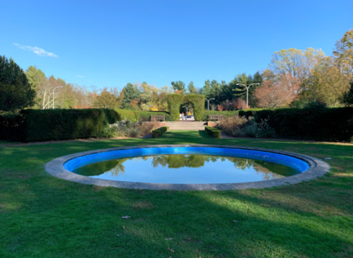 A circular pool reflects the blue sky, surrounded by lush greenery and neatly trimmed hedges in a serene garden setting.