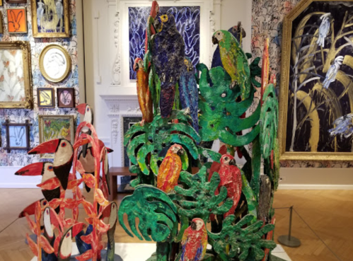 Colorful sculptures of birds and foliage displayed in an art gallery with patterned walls and framed artworks.