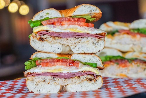 A stacked sandwich with layers of turkey, ham, lettuce, tomato, and egg on a toasted bagel, served on a checkered paper.