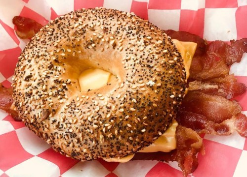 A toasted bagel with sesame seeds, melted cheese, and crispy bacon, served in a red and white checkered basket.