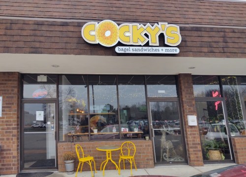 Exterior of Cocky's Bagel Sandwiches, featuring a sign, large windows, and a small outdoor seating area with yellow chairs.
