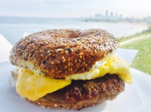 A sesame bagel sandwich with egg and sausage, set against a scenic waterfront view.