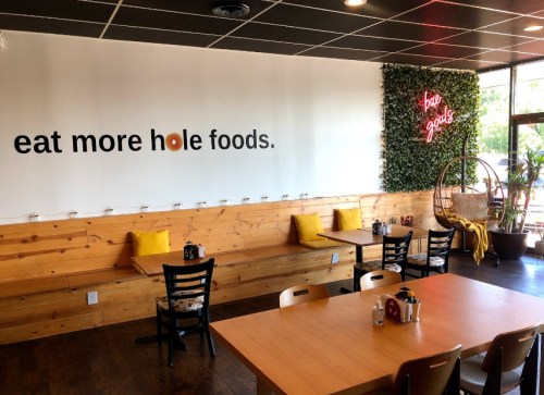 Interior of a cozy restaurant with wooden walls, tables, and a neon sign that says "eat more hole foods."