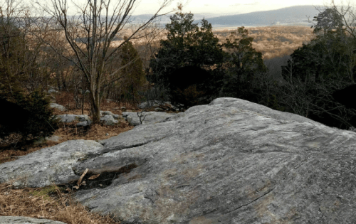 A rocky outcrop overlooks a valley with trees and distant hills under a cloudy sky.