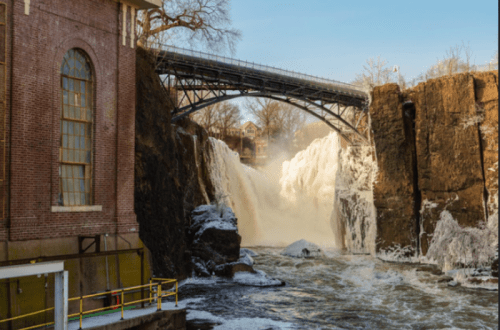 A waterfall cascades over rocky cliffs, with a bridge above and a brick building nearby, under a clear blue sky.