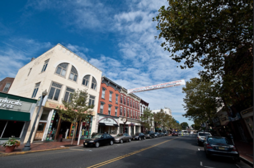 A charming street scene with shops, trees, and a blue sky, featuring a banner overhead and parked cars along the road.