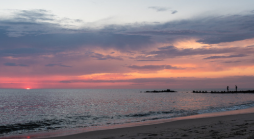A serene beach at sunset, with vibrant pink and orange skies reflecting on calm waters and a silhouette of a person on the shore.
