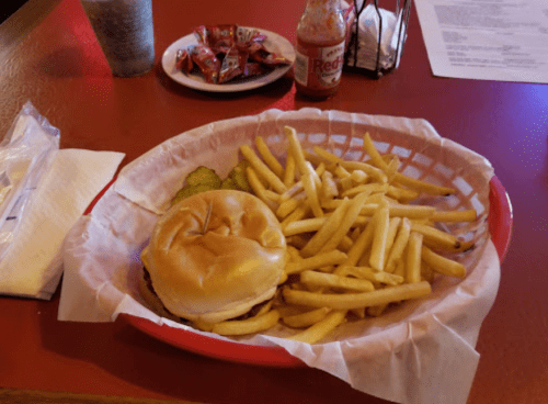 A burger with pickles and a side of golden fries in a red basket, with condiments and a drink in the background.