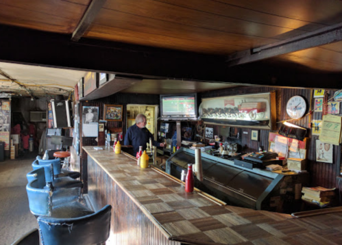 A rustic bar interior with a bartender, vintage decor, and condiments on the counter. A TV displays a sports game.