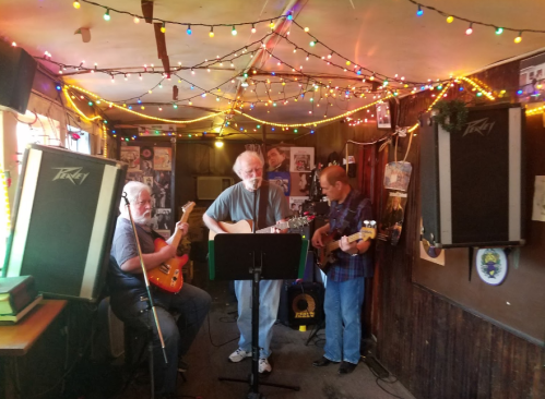 Three musicians perform in a cozy bar, surrounded by colorful string lights and posters, with amplifiers in the background.