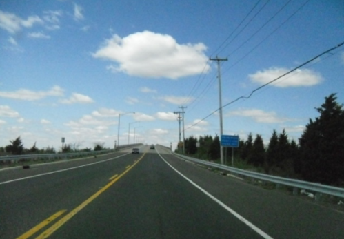 A winding road leads into the distance under a blue sky with fluffy clouds, flanked by trees and utility poles.