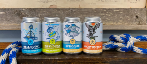 Four colorful beer cans lined up on a wooden surface, featuring nautical-themed designs and labels.