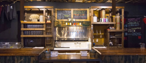 A rustic bar with wooden shelves, taps, and chalkboard menus, featuring a sink and various glassware.