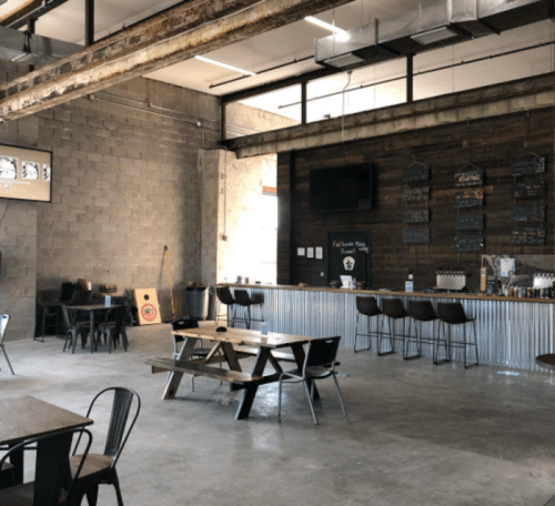 Interior of a modern, industrial-style bar with wooden tables, a metal bar, and a chalkboard menu on the wall.