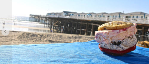 A colorful ice cream sandwich with a cookie on top, set against a beach and pier backdrop.