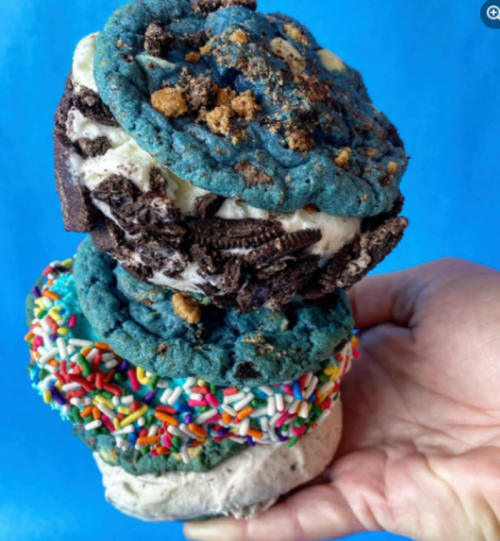 A hand holding three colorful ice cream sandwiches with blue cookies, sprinkles, and crushed cookies on top.