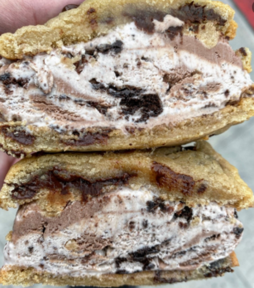 A close-up of a cookie ice cream sandwich filled with chocolate ice cream and cookie chunks, held in a hand.